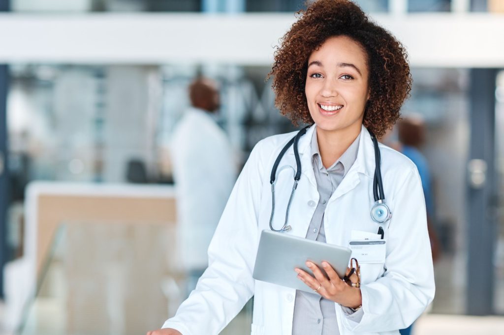 Confident female doctor with a stethoscope around her neck holding a tablet, smiling at the camera in a hospital setting.